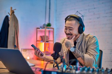 Asyalı bir adam kendi stüdyosunda podcast yaparken gülümsüyor..