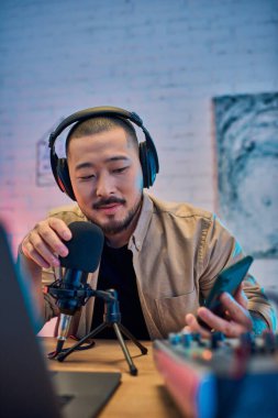 A handsome Asian man wearing headphones is recording a podcast in his home studio. clipart