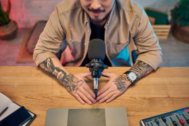 A tattooed Asian man sits in his podcast studio, preparing to record. clipart