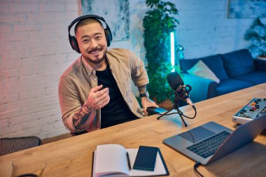 A smiling Asian man wearing headphones records a podcast in his studio. clipart