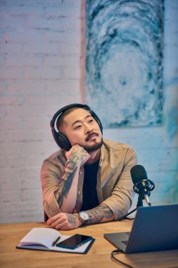 Stüdyo ortamında, kulaklık takmış, bronz gömlek giymiş yakışıklı bir Asyalı bir adam bir sonraki podcast bölümünü düşünüyor..
