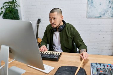 Asyalı bir adam stüdyosundaki bilgisayara bakarken MIDI klavye ve davul çalıyor..