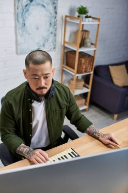 A handsome Asian man, wearing casual attire, sits at a desk in his studio, playing music on a keyboard and using a computer. clipart