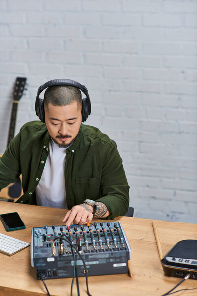 Красивый азиат в наушниках сидит за столом и смешивает музыку со звуковой панелью..