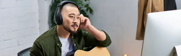 Stock image A handsome Asian man, wearing headphones, plays a guitar in his studio.