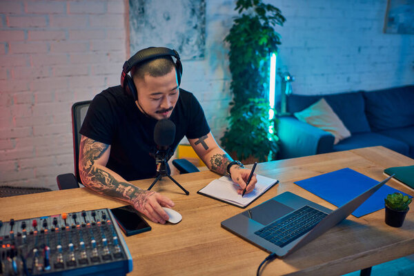 A young Asian man records a podcast in his studio, headphones on, microphone in hand.
