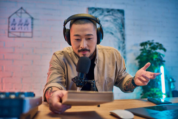 Красивый азиат записывает подкаст в своей студии, держит книгу и говорит в микрофон.