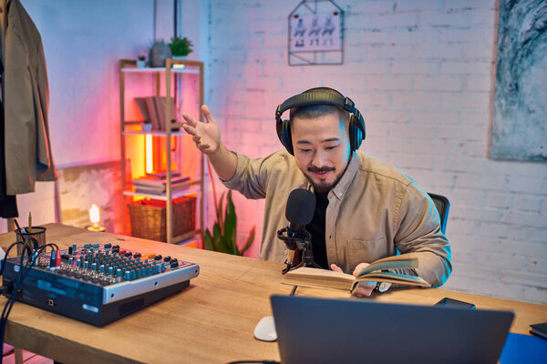 An Asian man records a podcast in his home studio, with a microphone, headphones, and a book.