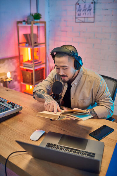 Красивый азиат в наушниках сидит за столом в своей студии, читает из книги во время записи подкаста.