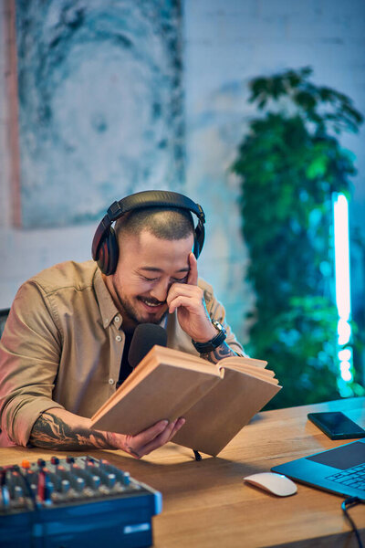 An Asian man with headphones smiles while reading from a book in his podcast studio.