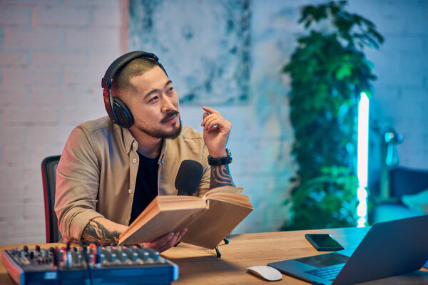 A handsome Asian man sits in his podcasting studio, wearing headphones and holding a book.