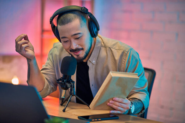 Красивый азиат записывает подкаст в своей студии, держит книгу и говорит в микрофон.