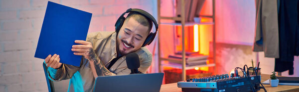 A young Asian man with headphones, microphone, and a blue folder, smiling and recording in a home studio.
