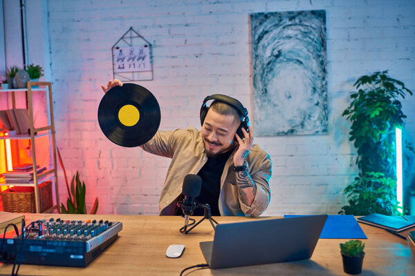 Красивый азиат подкастинг в студии, держащий виниловую пластинку и улыбающийся.