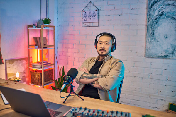 A handsome Asian man wearing headphones sits in his podcast studio, preparing to record.