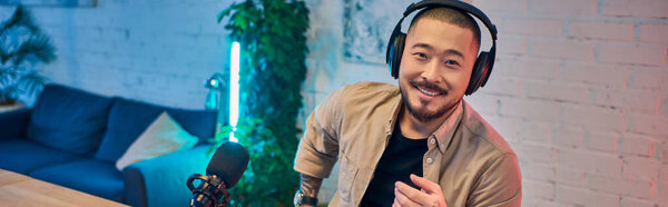A handsome Asian man with headphones on smiles as he records a podcast in his home studio.
