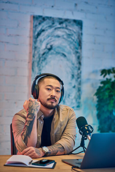 Татуированный азиат в наушниках сидит в своей студии, задумчиво глядя в камеру во время записи подкаста.