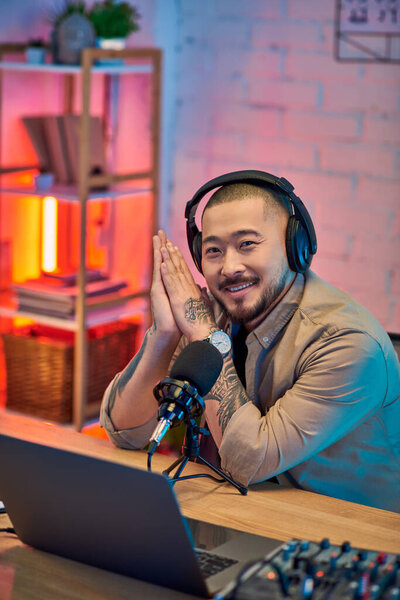 A handsome Asian man wearing headphones smiles while podcasting in his home studio.