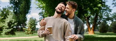 İki sakallı eşcinsel, günlük kıyafetleriyle yeşil bir parkta gülüyorlar, neşe ve sevgi dolu bir anı paylaşıyorlar..