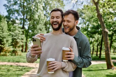 A bearded gay couple shares a laugh while looking at a phone and enjoying coffee in a green park. clipart