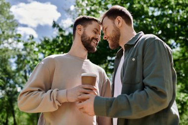 Two bearded men share a coffee and a loving moment in a green park on a sunny day. clipart