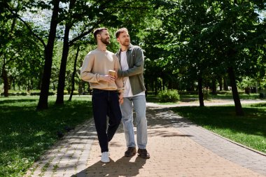 Two bearded men, dressed casually, hold hands and walk through a park on a sunny day. clipart