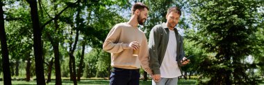 Two bearded men, dressed casually, walk hand-in-hand in a green park on a sunny day. clipart