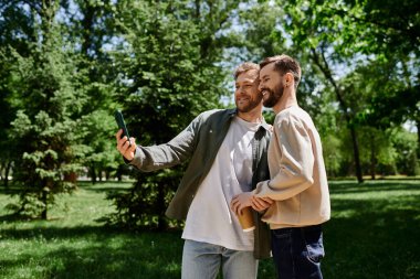 A bearded gay couple takes a selfie in a green park, enjoying a sunny afternoon. clipart