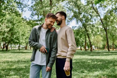 Two bearded men in casual clothing laugh and enjoy each others company in a green park. clipart