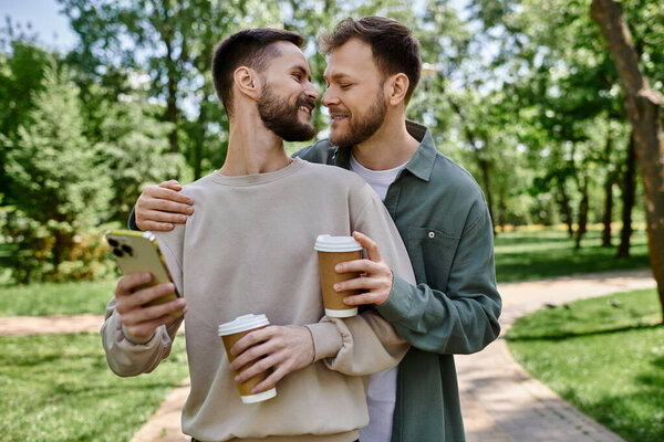 Бородатая гей-пара гуляет в парке, держа в руках кофе и телефон, с любящим взглядом, разделенным между ними.