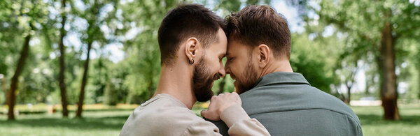 Two bearded gay men in casual attire stand close together in a green park.