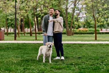 Sıradan giyinmiş sakallı eşcinsel bir çift gülümsüyor ve labrador köpeğiyle parkta bir günün tadını çıkarıyor..