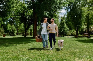 Sakallı eşcinsel bir çift, güneşli bir öğleden sonranın tadını çıkarırken yeşil bir parkta el ele yürür..