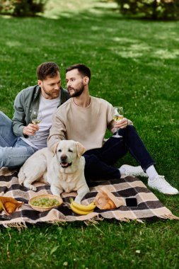 İki sakallı adam yeşil bir parkta labrador köpeğiyle pikniğin tadını çıkarıyor..