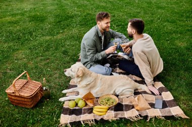 Sakallı eşcinsel bir çift, labrador av köpeğiyle çimenli bir parkta pikniğin tadını çıkarıyor..