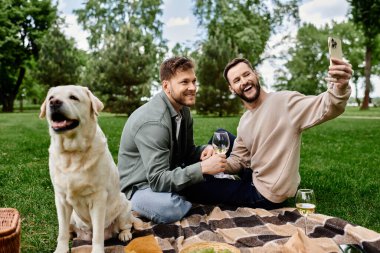 A bearded gay couple enjoys a picnic in a green park with their labrador retriever. One partner takes a selfie while the other smiles. clipart