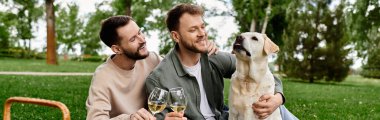 İki sakallı adam yeşil bir parkta Labrador av köpeğiyle pikniğin tadını çıkarıyor..