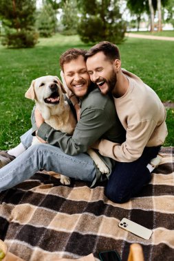 İki sakallı adam ve onların Labrador köpeği yeşil bir parkta piknik yaparken keyifli bir anı paylaşıyorlar..