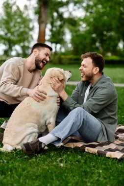 Sakallı eşcinsel bir çift yeşil bir parkta piknik yaparken labrador köpeğiyle neşeli bir anı paylaşıyor..