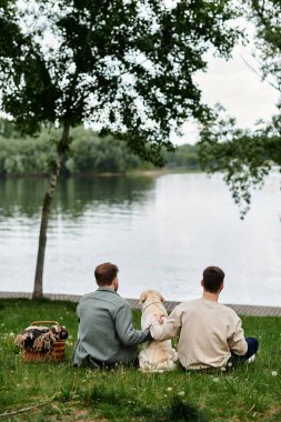 A bearded gay couple enjoys a picnic with their labrador dog in a lush green park by the river. clipart