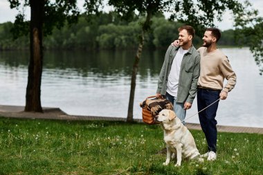 Sakallı eşcinsel bir çift göl kenarındaki yeşil bir parkta piknik yaparken labrador köpeğiyle sevgi dolu bir anı paylaşıyor..