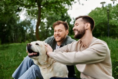 Labrador köpeğiyle mutlu bir eşcinsel çift yemyeşil bir parkta güneşli bir öğleden sonranın tadını çıkarıyor..