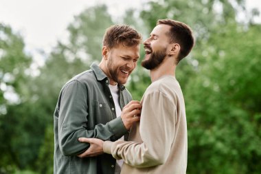 Sakallı bir LGBTQ çifti, yeşil bir parkta güneşli bir günün tadını çıkarırken aynı kahkahayı paylaşıyor..