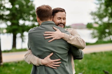 Two bearded men embrace in a loving hug while enjoying a sunny day in a beautiful park. clipart