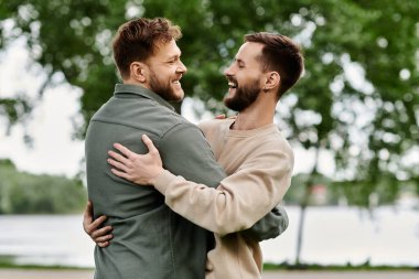 Two bearded men embrace in a park, laughing and looking at each other with love. clipart