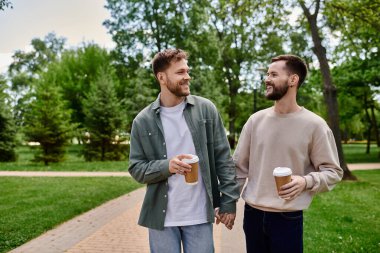 A bearded gay couple walks hand-in-hand through a park, enjoying a coffee together. clipart