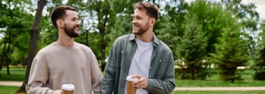 Two bearded men, dressed casually, walk together in a park, smiling and holding coffee cups. clipart