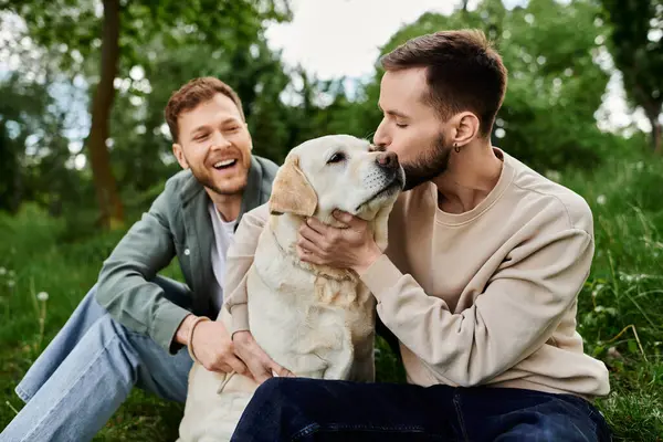 İki sakallı adam, çimenli bir parkta Labrador köpeğiyle eğleniyor. Bir adam köpeği öpüyor..