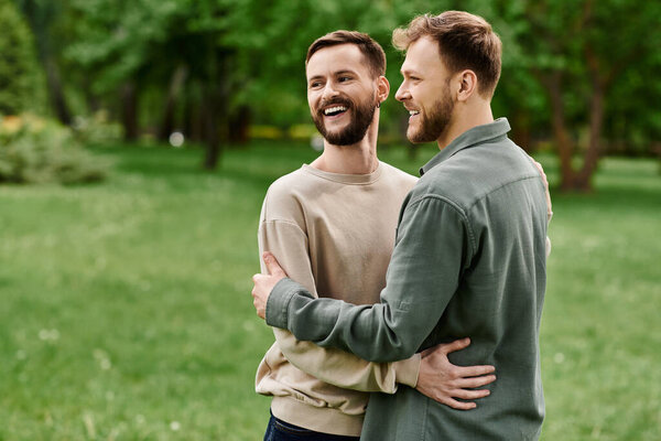 Two bearded men embrace each other while laughing in a grassy park.