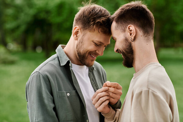 Two bearded gay men stand close together in a green park, their heads touching as they share a tender moment.